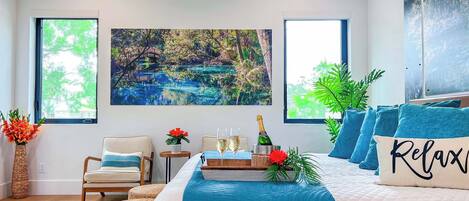 Um quarto com uma cama grande, uma mesinha com copos e uma garrafa, e um quadro de uma paisagem.