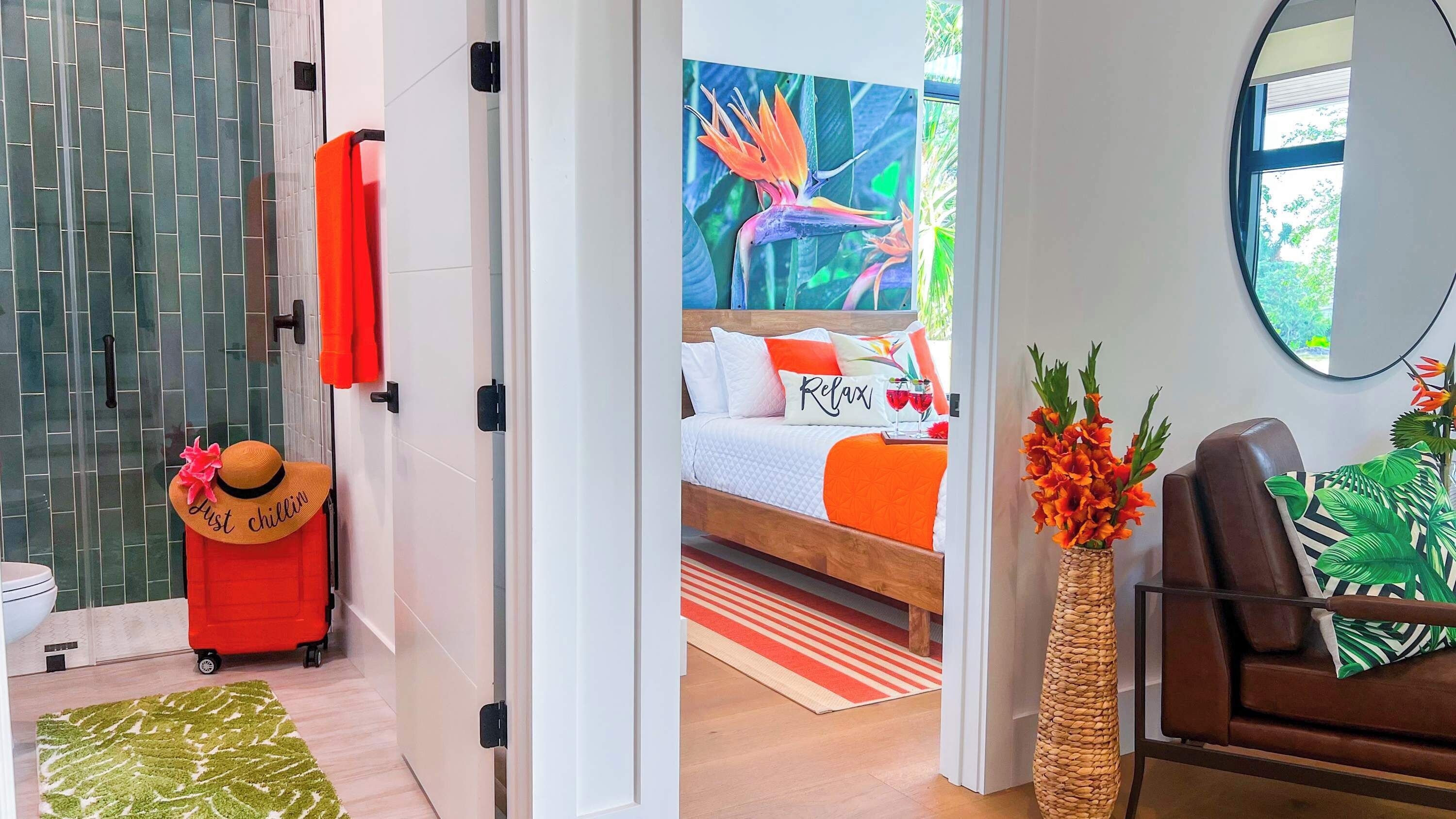 Une chambre d’hôtel avec un lit, une valise rouge, un chapeau et un tableau sur le thème des pays tropicaux.