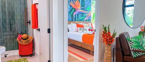 Une chambre d’hôtel avec un lit, une valise rouge, un chapeau et un tableau sur le thème des pays tropicaux.