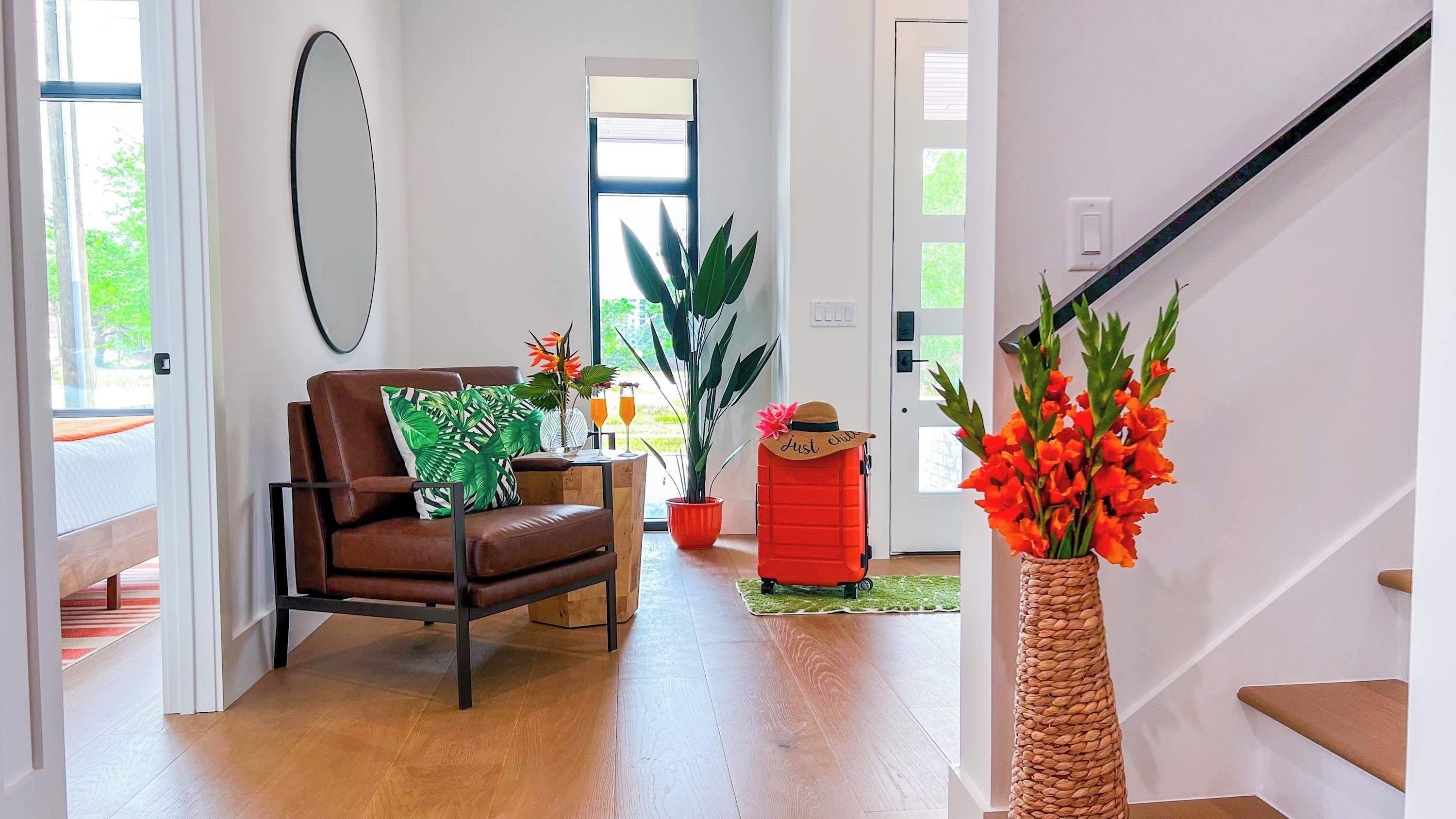A modern living room with a brown leather chair, a red ottoman, a large potted plant, and a tall woven vase with orange flowers.