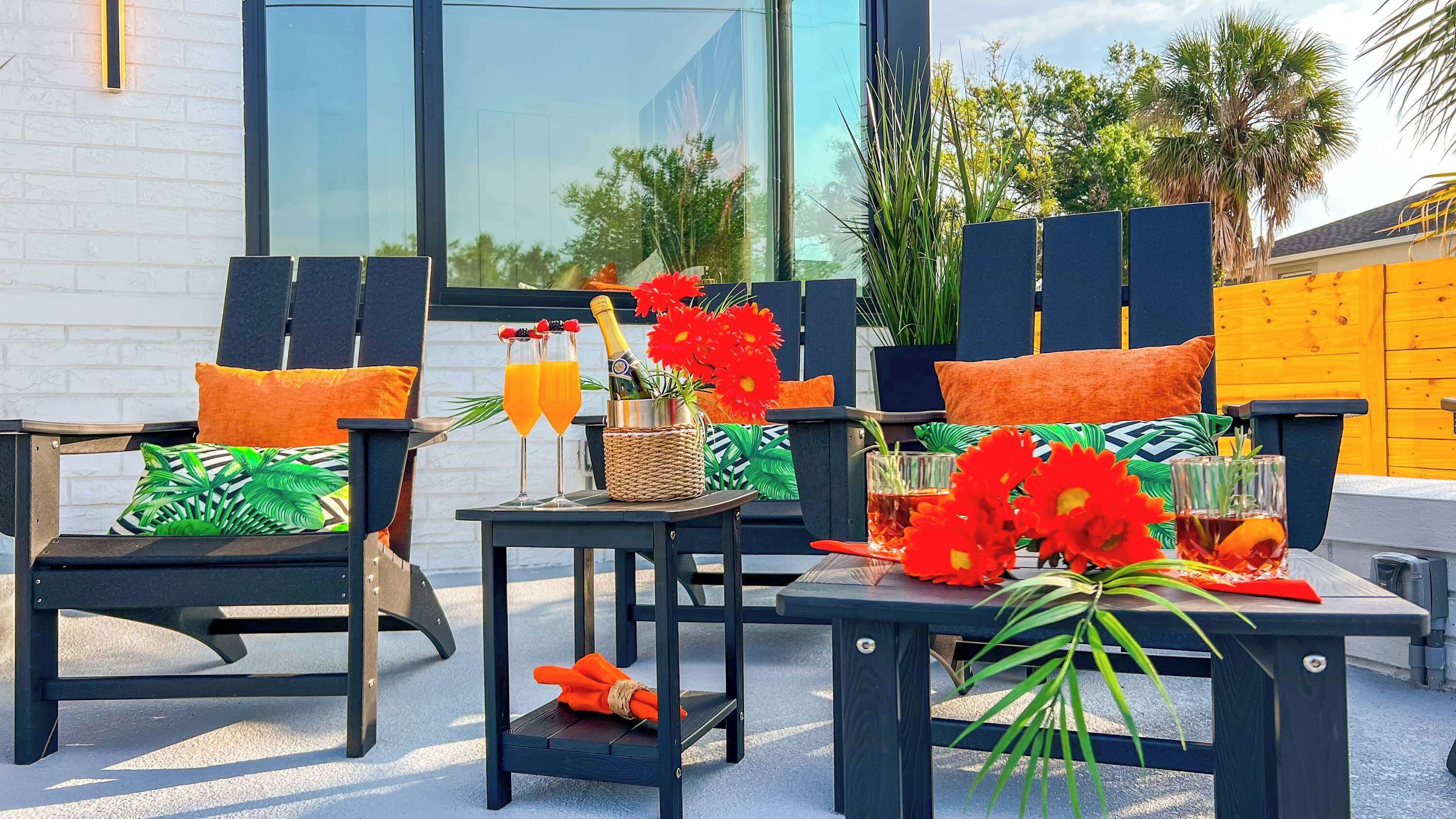 Un patio con mobiliario exterior, incluyendo dos sillas con cojines, una mesa pequeña con bebidas y flores, y un fondo con un edificio y vegetación.