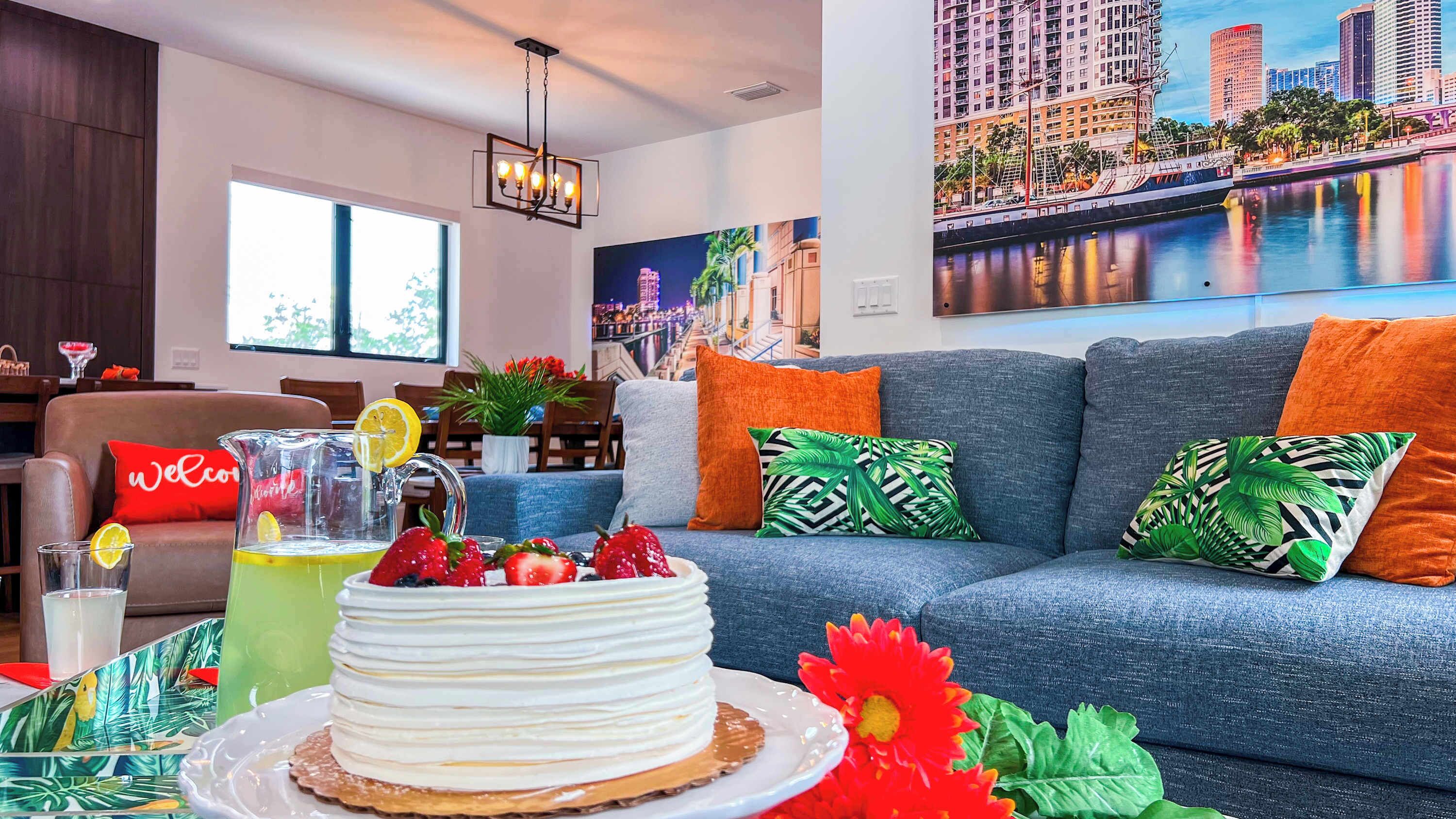 Un salon avec un canapé, une table sur laquelle se trouvent un gâteau et des boissons, et un tableau représentant un paysage urbain accroché au mur.