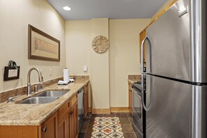 A compact kitchen with a stainless steel refrigerator, double sink, wooden cabinets, and a decorative tile floor.
