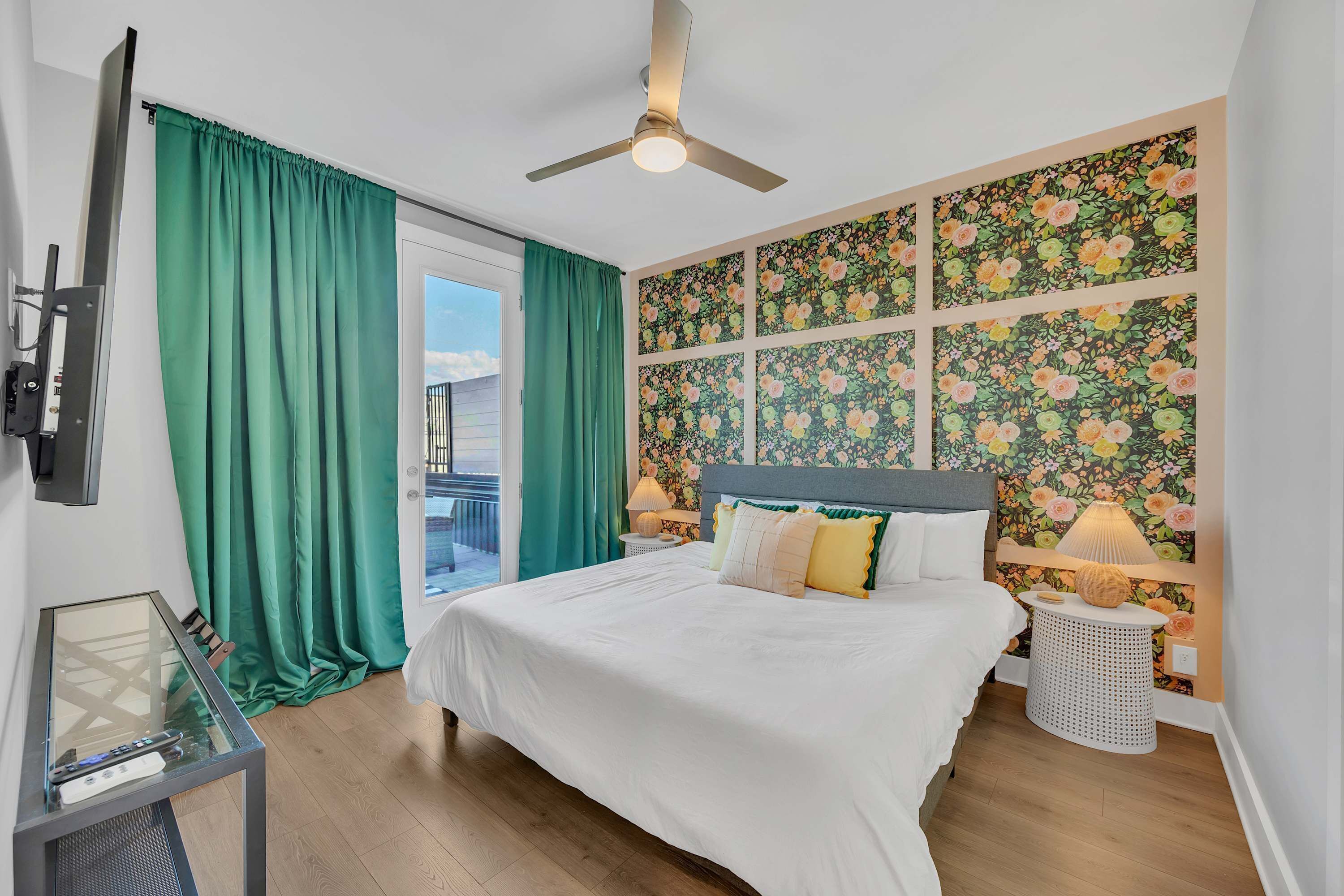 Une chambre à coucher comprenant un lit, un téléviseur, un ventilateur de plafond, une fenêtre avec des rideaux, une table de chevet avec une lampe et un mur recouvert d’un papier peint à motifs floraux.