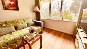 Un salón con un sofá floral, una mesa de centro de vidrio, una lámpara y vistas a árboles a través de amplios ventanales.