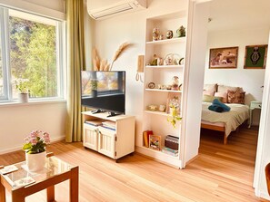 A room with a television on a white stand, a bookshelf with decorative items, a bed with pillows, and a window with a view of trees.