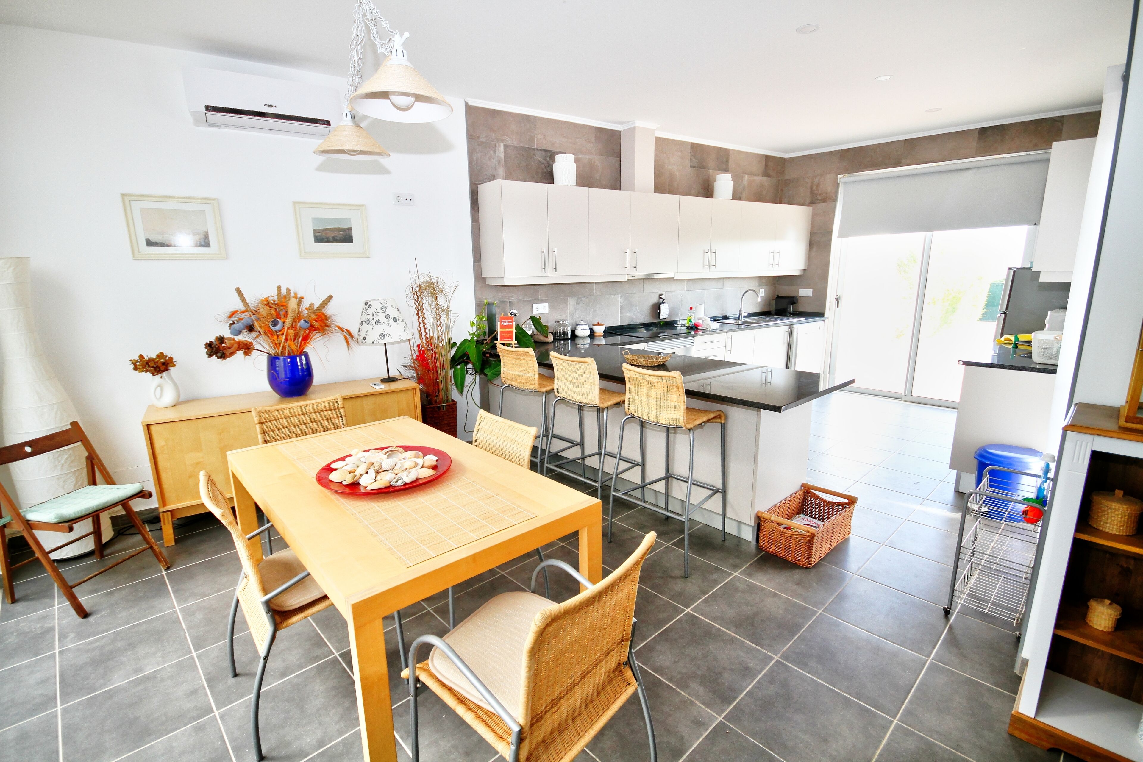 A modern kitchen with white cabinets, a dining area with a wooden table and wicker chairs, and a large window providing natural light.
