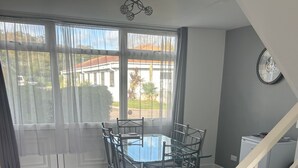 A dining area with a glass-top table and metal chairs, a staircase, and a view of a residential area through large windows. - Modern, 2BR Villa Walk to Beach Parking & Wifi Dawlish Warren (Dawlish)
