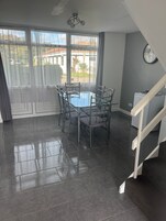 Un comedor con una mesa de vidrio, sillas de metal, una escalera y vistas a una zona residencial a través de amplios ventanales.