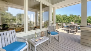 A covered outdoor deck with wooden furniture, including a table and chairs, and a large window offering a view of greenery. - Oceanfront, private boardwalk and pool (Kiawah Island)