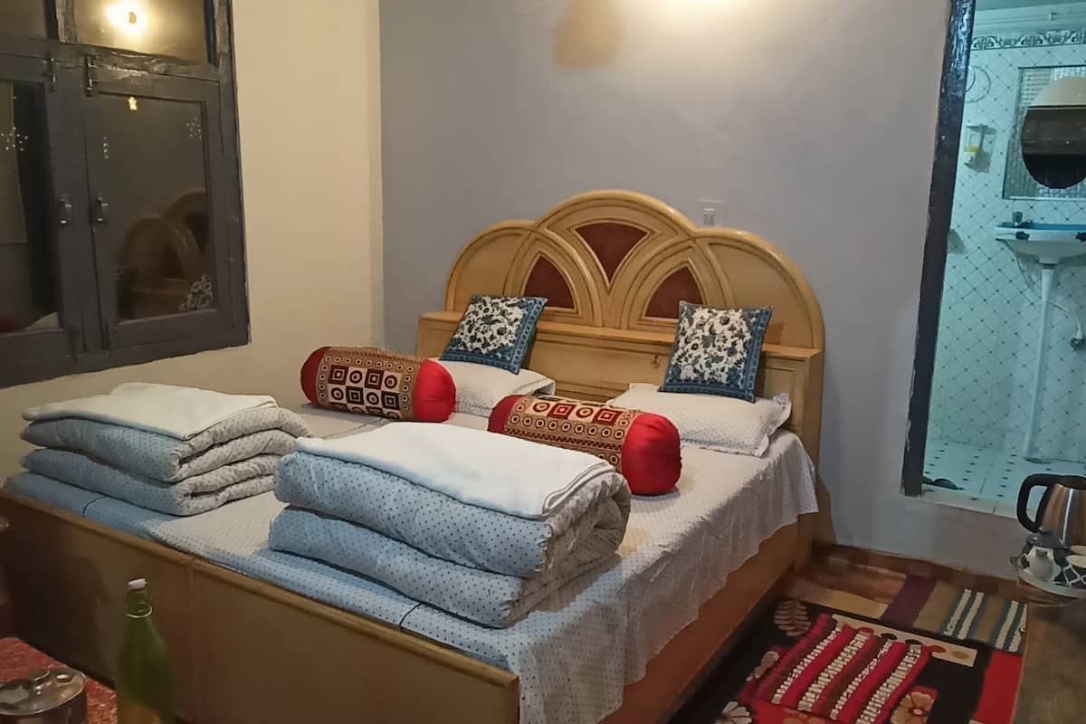 A bedroom with a wooden ceiling, a bed with patterned pillows, a nightstand, a table with a bottle, and a window with a view of a sink.