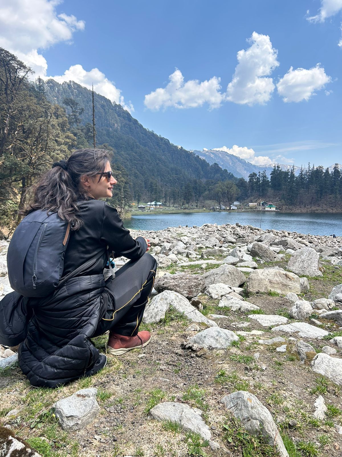 A person sitting on a rocky shore by a lake with mountains in the background.