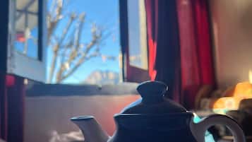 Una mesa con una tetera y una taza, una ventana que da a las copas de los árboles, y una cortina roja.