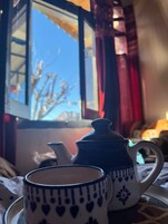 Una mesa con una tetera y una taza, una ventana que da a las copas de los árboles, y una cortina roja.
