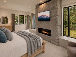 Une chambre avec une cheminée en pierre, un lit recouvert d’une couverture, un téléviseur fixé au mur et une vue sur la verdure à l’extérieur.