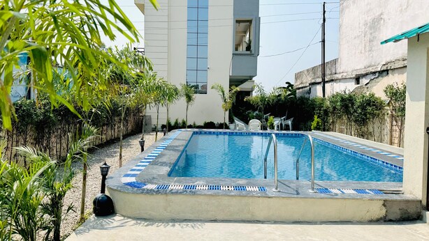 A rectangular swimming pool with blue tiles and a white border, surrounded by small trees and a concrete deck. - Corbett Hideaway Dada 'S (Ramnagar)