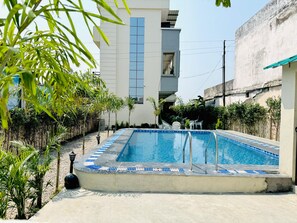 A rectangular swimming pool with blue tiles and a white border, surrounded by small trees and a concrete deck. - Corbett Hideaway Dada 'S (Ramnagar)