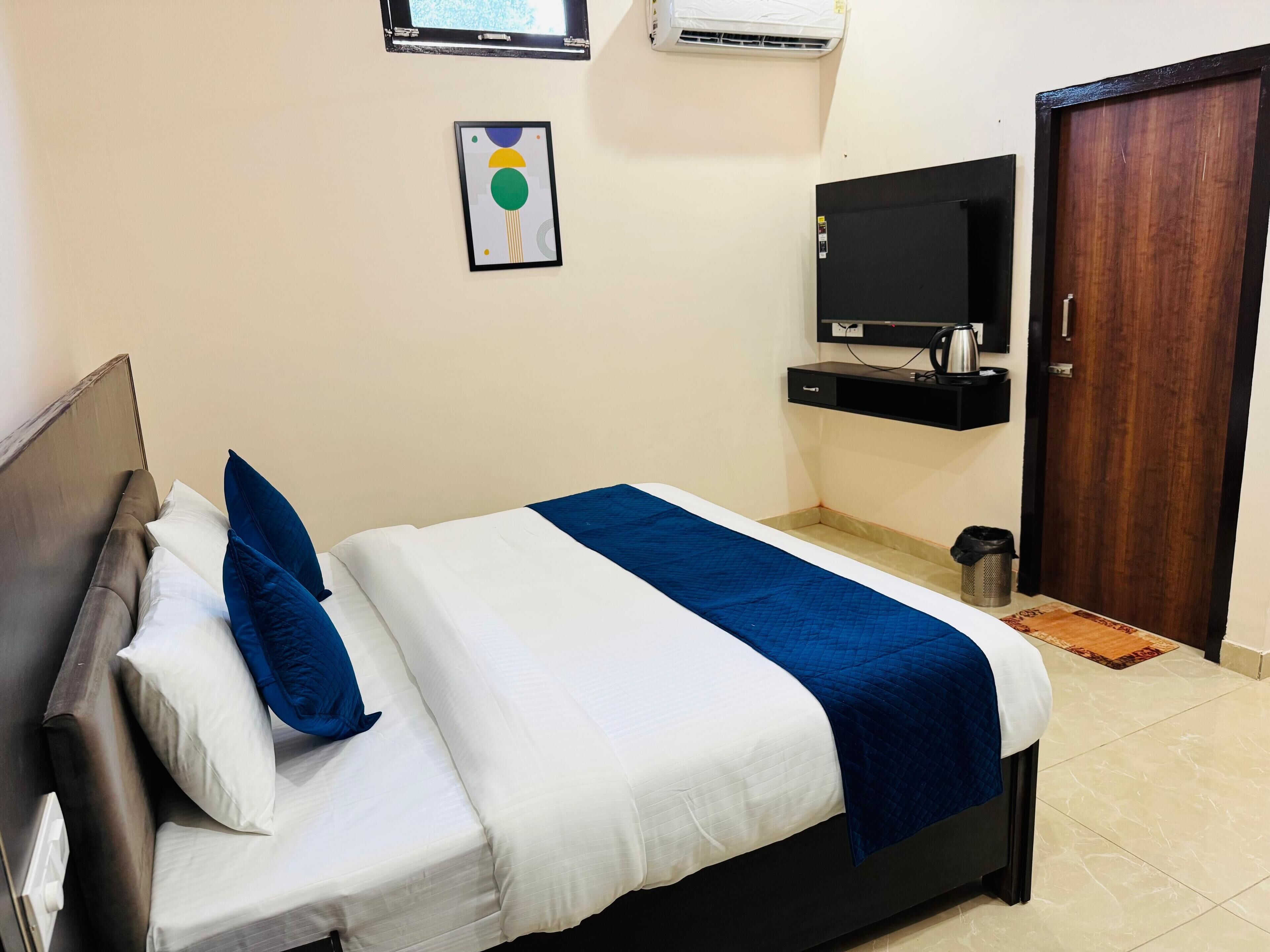 A neatly made bed with white and blue bedding, a television mounted on the wall, a wooden door, and a small trash bin.