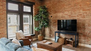 Ein Wohnzimmer mit Backsteinwand, großem Fenster, einer Couch, einem Couchtisch, einem Fernseher auf einem Schrank und einem Topf mit Pflanze.