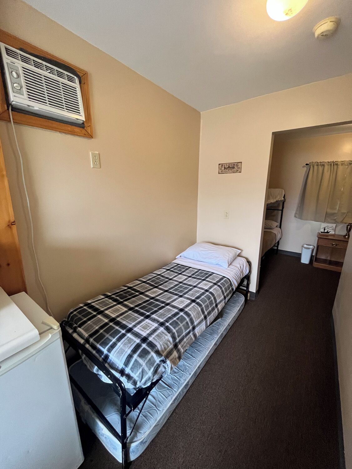 Quarto pequeno com uma cama de solteiro, colcha xadrez, aquecedor fixado na parede, mesa de cabeceira e janela com cortinas.