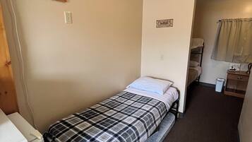 Quarto pequeno com uma cama de solteiro, colcha xadrez, aquecedor fixado na parede, mesa de cabeceira e janela com cortinas.