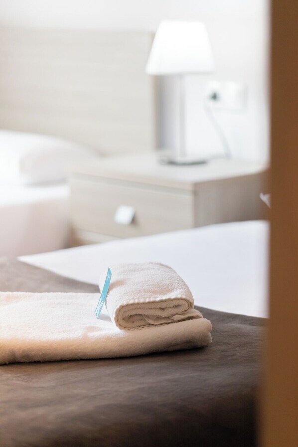 Un lit bien fait, avec une serviette blanche pliée posée sur une surface en bois, une lampe de chevet et une table de chevet blanche, dans une chambre d’hôtel.