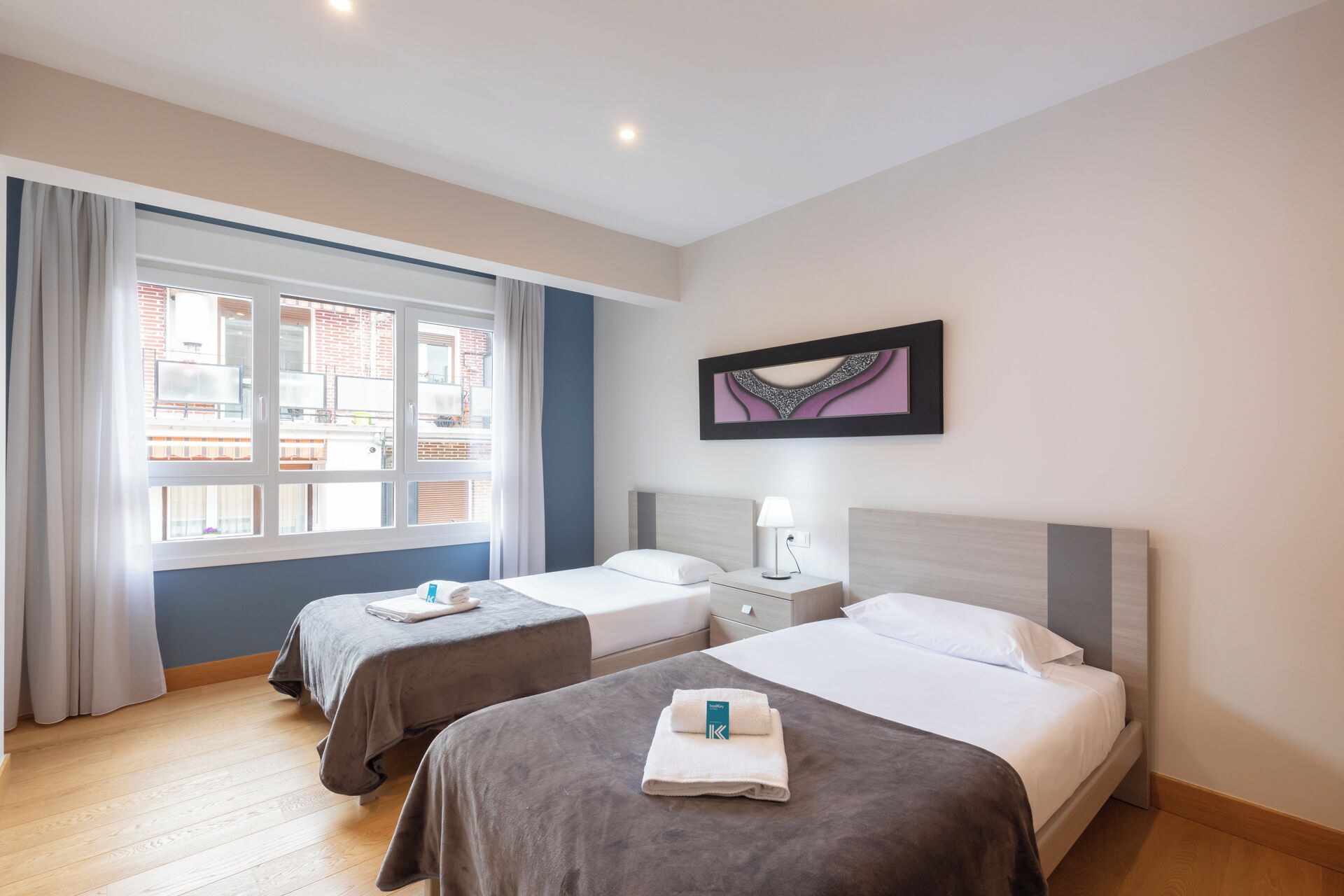 A hotel room with two beds, a window with curtains, a bedside table with a lamp, and a framed artwork on the wall.