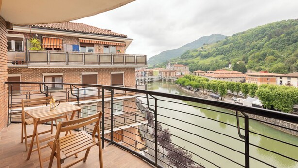 Un balcón con mobiliario de madera, vista a un río y edificios al fondo.