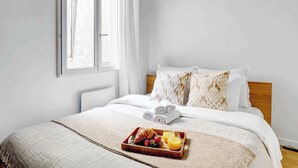 Un lit bien fait, avec des serviettes pliées, un plateau contenant des croissants, des fraises et du jus d’orange, et une fenêtre avec des rideaux.