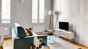 Un salon moderne avec un canapé de couleur turquoise, un meuble TV blanc, une lampe sur pied et une grande fenêtre.