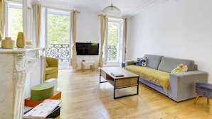 Un salón espacioso con un sofá gris, una mesa de centro y un televisor. Hay ventanas amplias con cortinas, un balcón y una chimenea con adornos decorativos.
