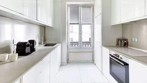 Una cocina moderna con armarios blancos, una cafetera negra y un ventanal con contraventanas blancas.