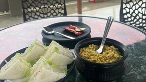 Una mesa con una comida que incluye sándwiches y un tazón de fideos, ubicada en un balcón con vistas a las montañas.