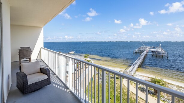 A balcony with a wicker chair and a small table, overlooking a beach and a pier with boats. - What A View! Marina, Intracoastal, Gulf Islands National Seashore & The Gulf (Pensacola)