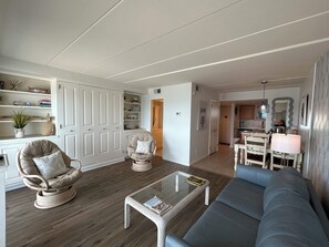 Una sala de estar con un sofá, una mesa de centro de vidrio, una silla de mimbre, una zona de comedor con una mesa y sillas, y una cocina al fondo.
