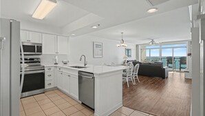 A modern kitchen with white cabinetry, a built-in oven, and a dishwasher. There is a dining area with a table and chairs, and a balcony with a view of the ocean. - Enjoy a memorable beach vacation! | Laguna Keyes -  903 (North Myrtle Beach)
