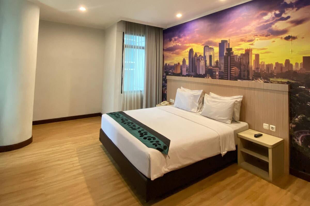 A hotel room with a bed, a nightstand, a cityscape mural, and a window with curtains.