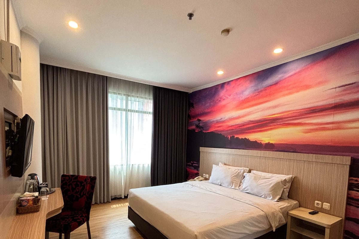 A hotel room with a large bed, a desk, a chair, a nightstand, and a wall mural.