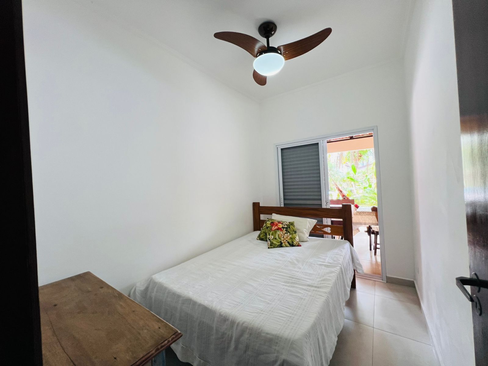 A small, clean room with a single bed, a ceiling fan, and a window with a view of greenery outside.