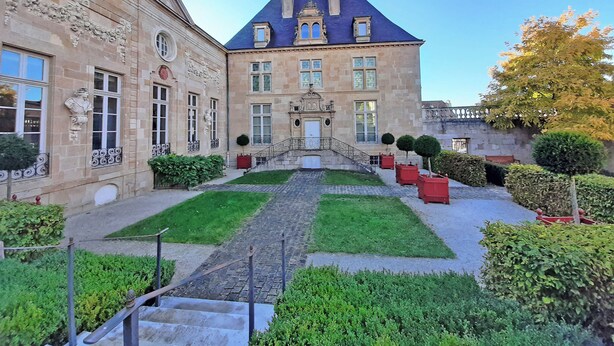 A historic building with a symmetrical facade, a central entrance, and a well-maintained garden with hedges and potted plants.