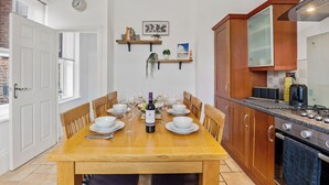 Een eetgedeelte met een houten tafel, gedekt voor zes personen, omringd door stoelen. Op de tafel staan een fles wijn en glazen. De kamer heeft houten kasten, een fornuis en een klok aan de muur.