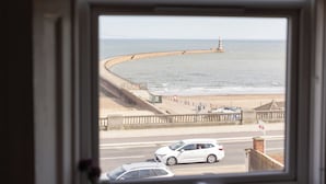 Uitzicht op de kust door een raam, met een zeedijk, strand en een vuurtoren.