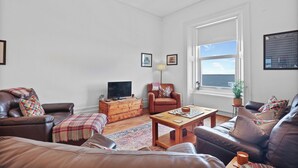 Een woonkamer met een bruine leren bank, een houten salontafel, een rode fauteuil en een televisie op een houten dressoir.