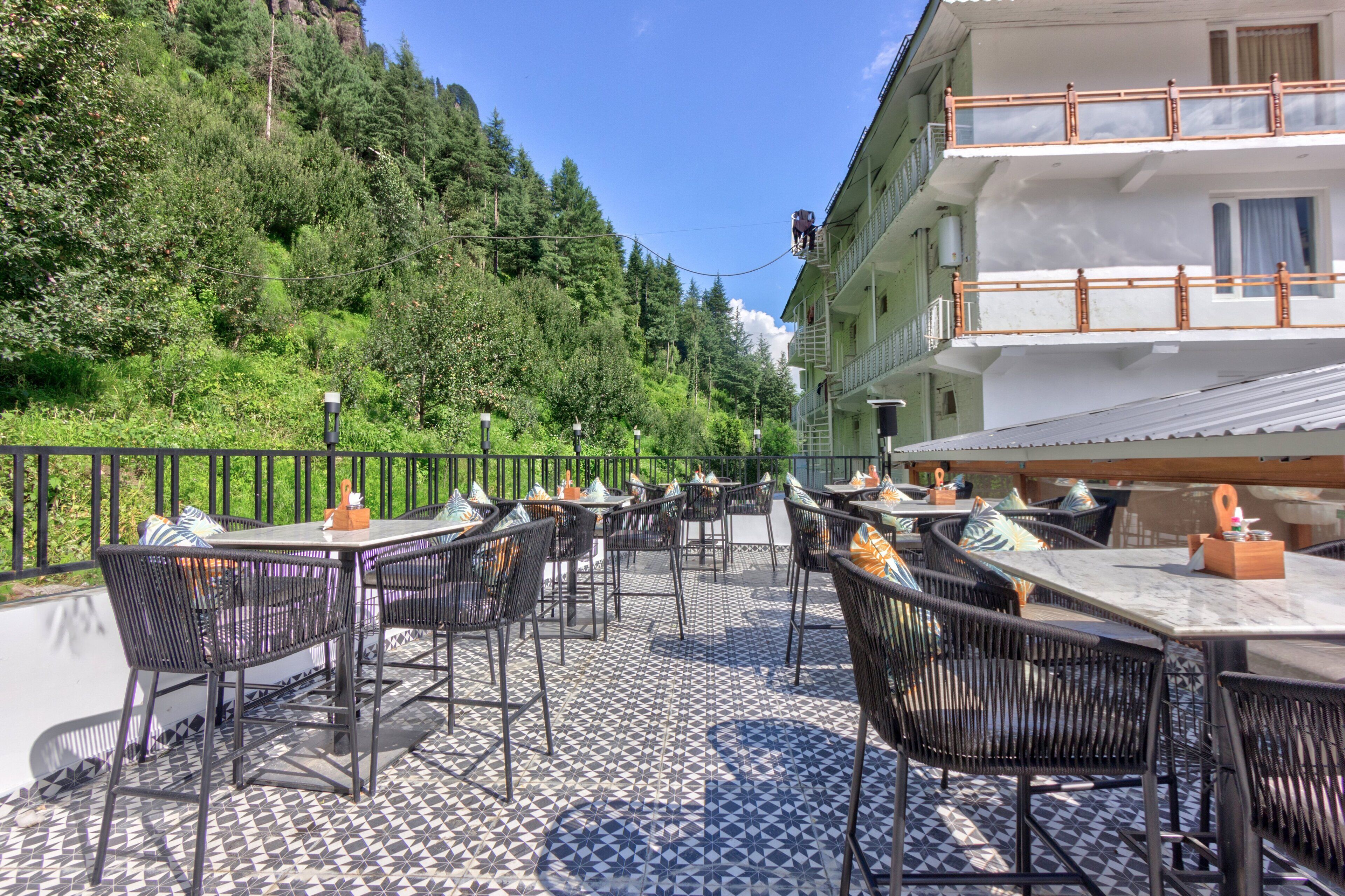 An outdoor patio with tables and chairs, surrounded by a railing, overlooking a forested hillside.