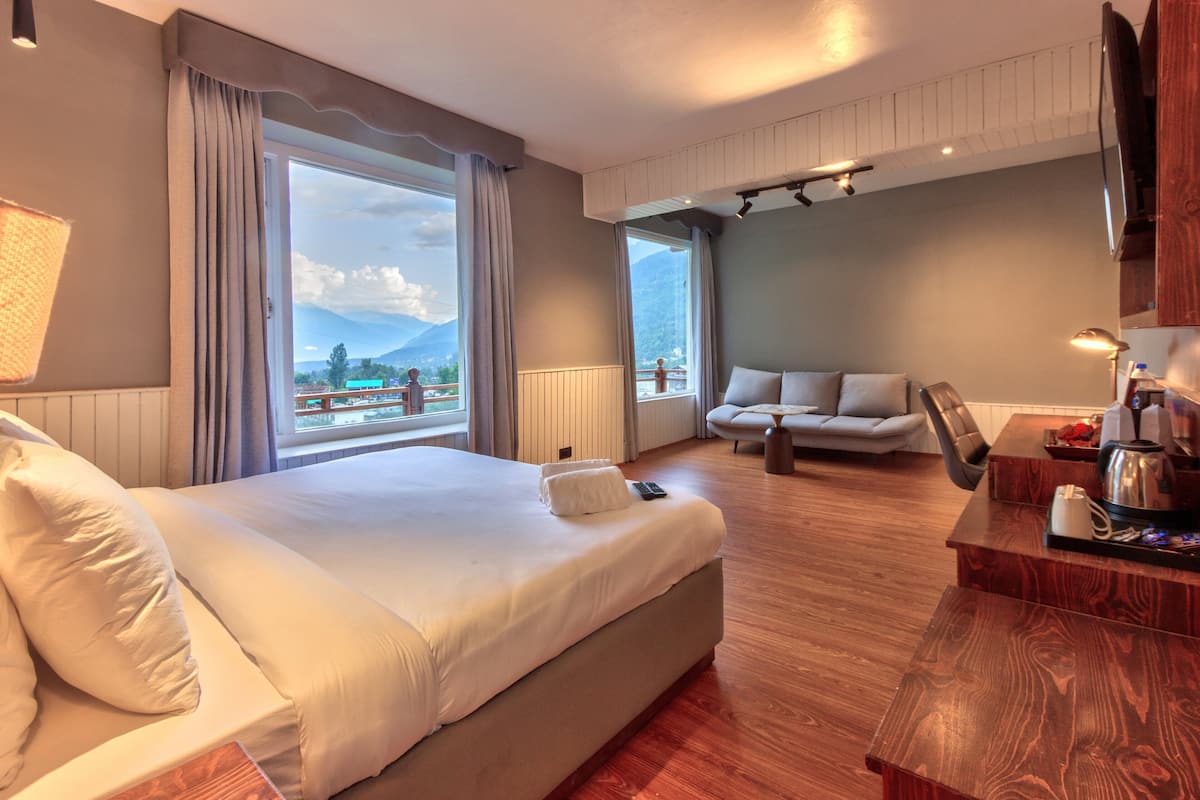 Habitación de hotel con una cama grande, un sofá, una mesita de madera y vistas a montañas desde la ventana.