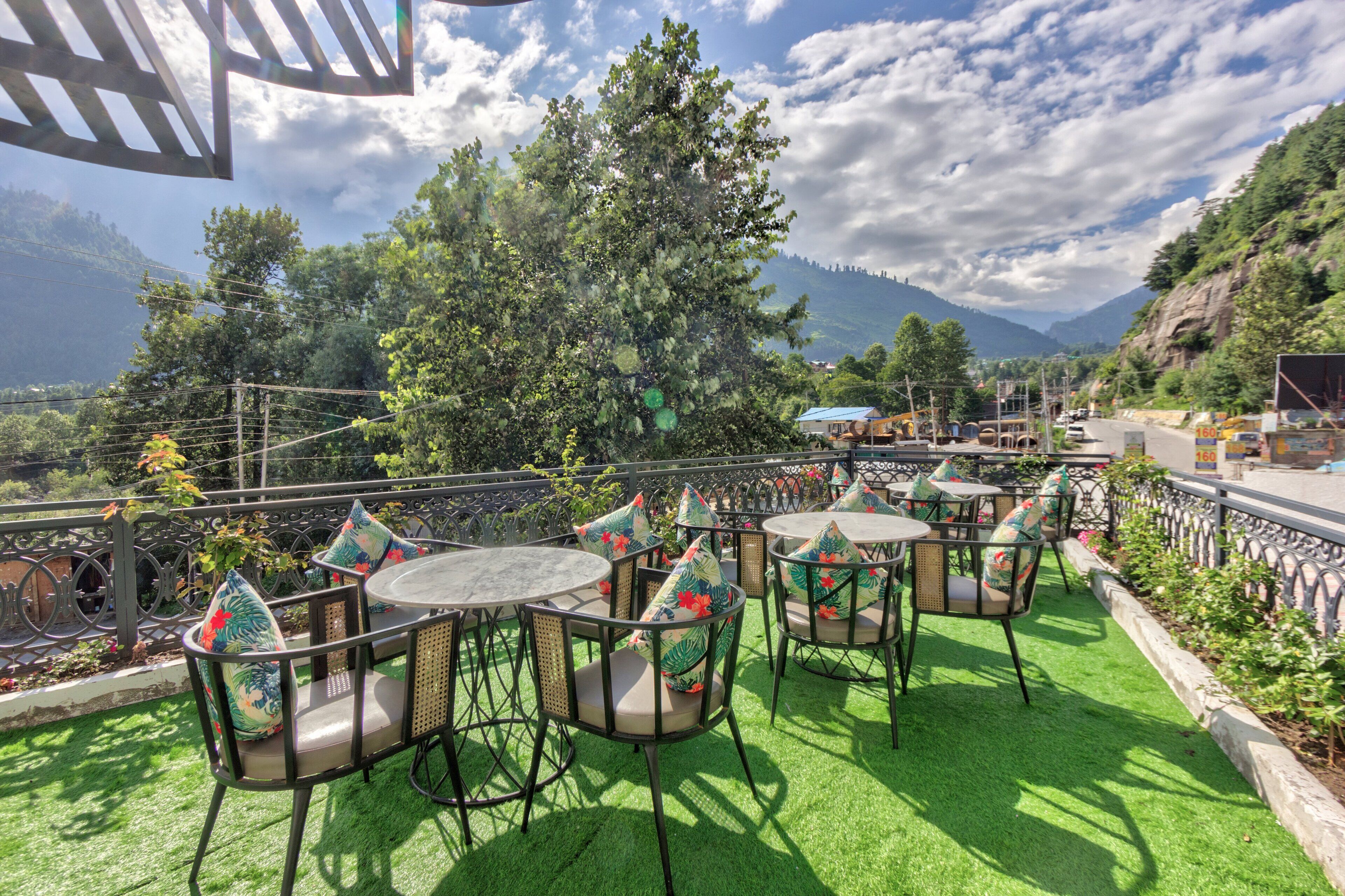 An outdoor seating area with round tables and chairs, surrounded by greenery and a mountainous backdrop.