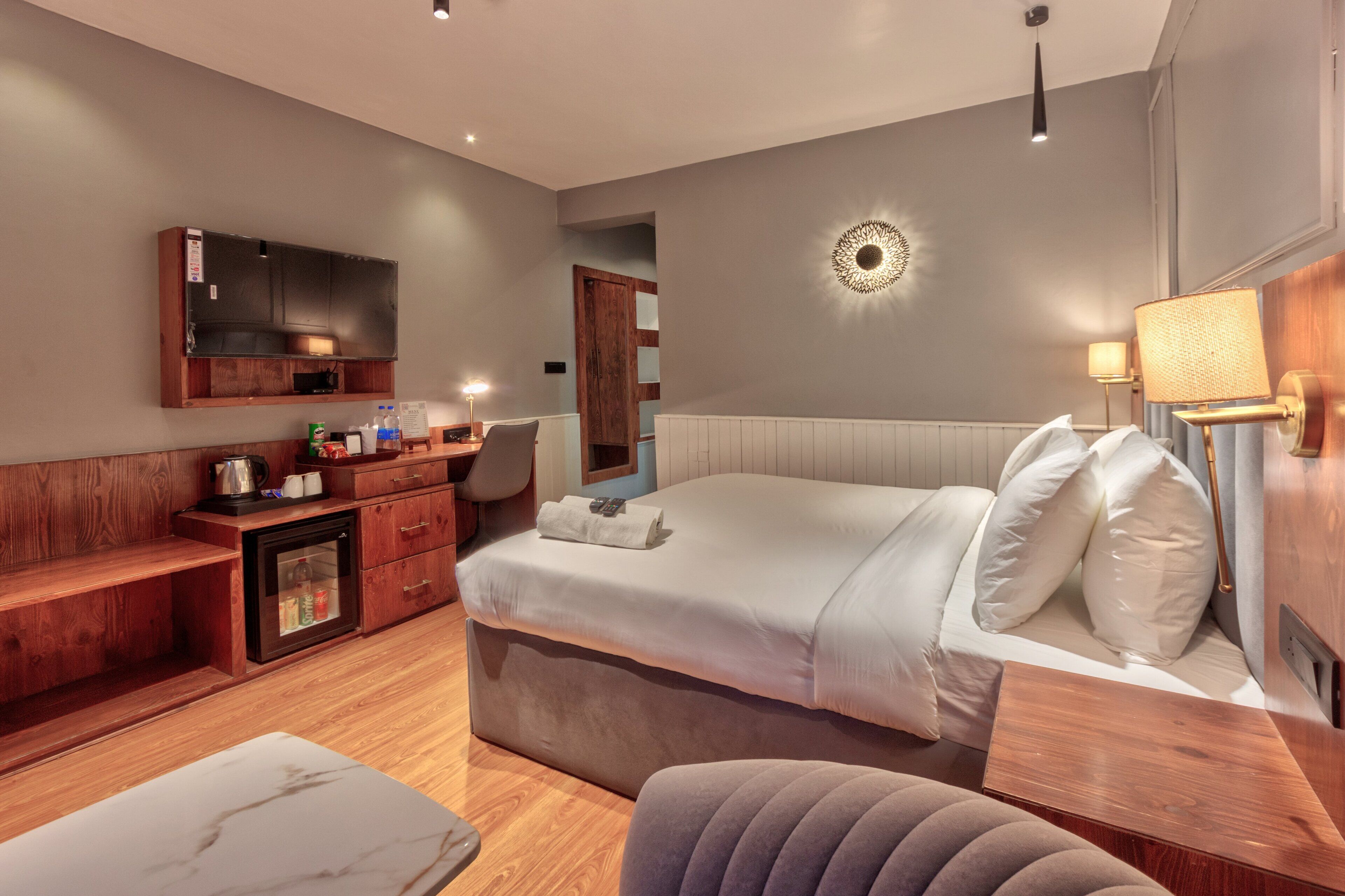 A modern hotel room with a large bed, a wooden dresser, a small kitchen area, and a wall-mounted TV.