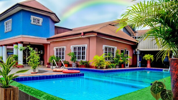 Ein Swimmingpool mit blaugrünem Rand, umgeben von Kübelpflanzen und einem Haus mit rotem Dach.