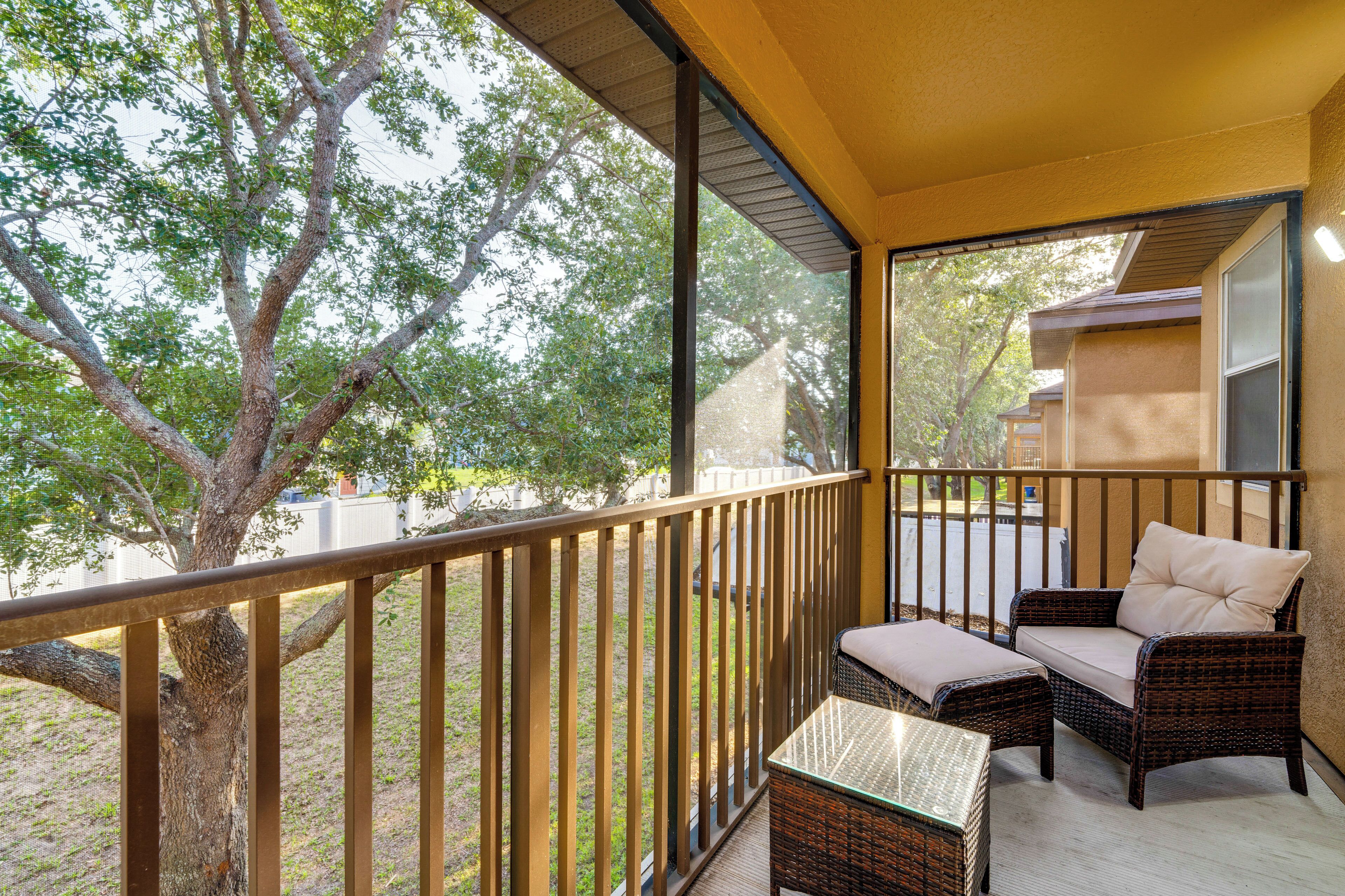A balcony with wicker furniture, a glass table, and a view of trees and a building.