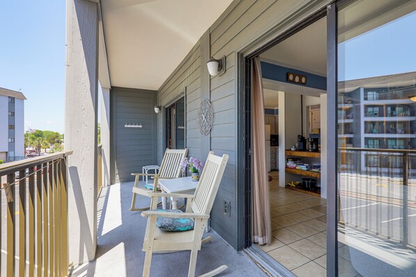 A balcony with two rocking chairs and a small table, a sliding glass door leading to an interior space with shelves and a kitchen area. - Steps to Shore: Family Retreat in Myrtle Beach (Myrtle Beach)